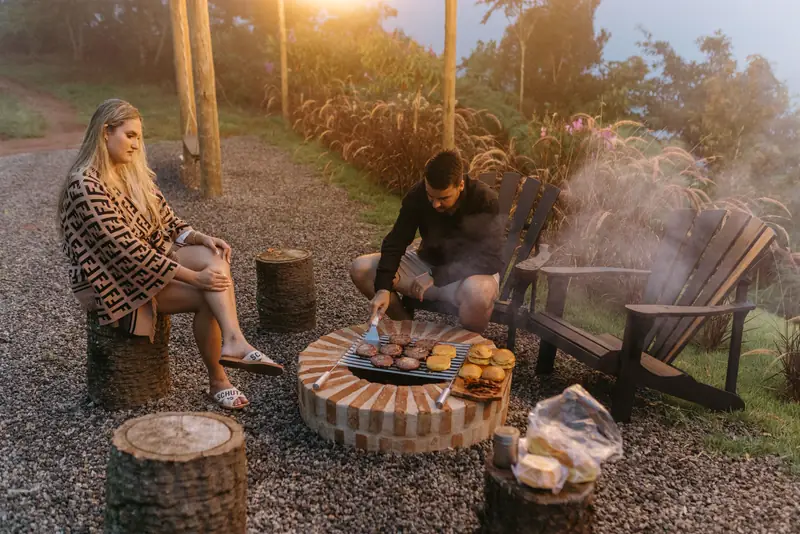 Two friends at dusk by a round stone fire pit with log-round seats, one tending the fire